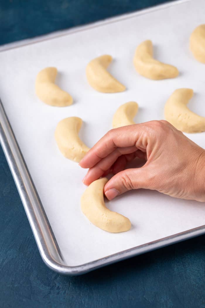 Hand forming almond crescent cookie dough into a crescent shape on a parchment-lined baking sheet.