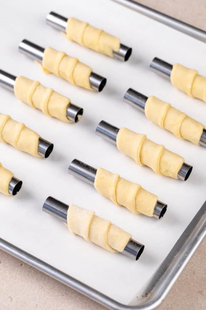 Strips of clothespin cookie dough wrapped around mini cannoli forms, set on a parchment-lined baking sheet, ready to go in the oven.