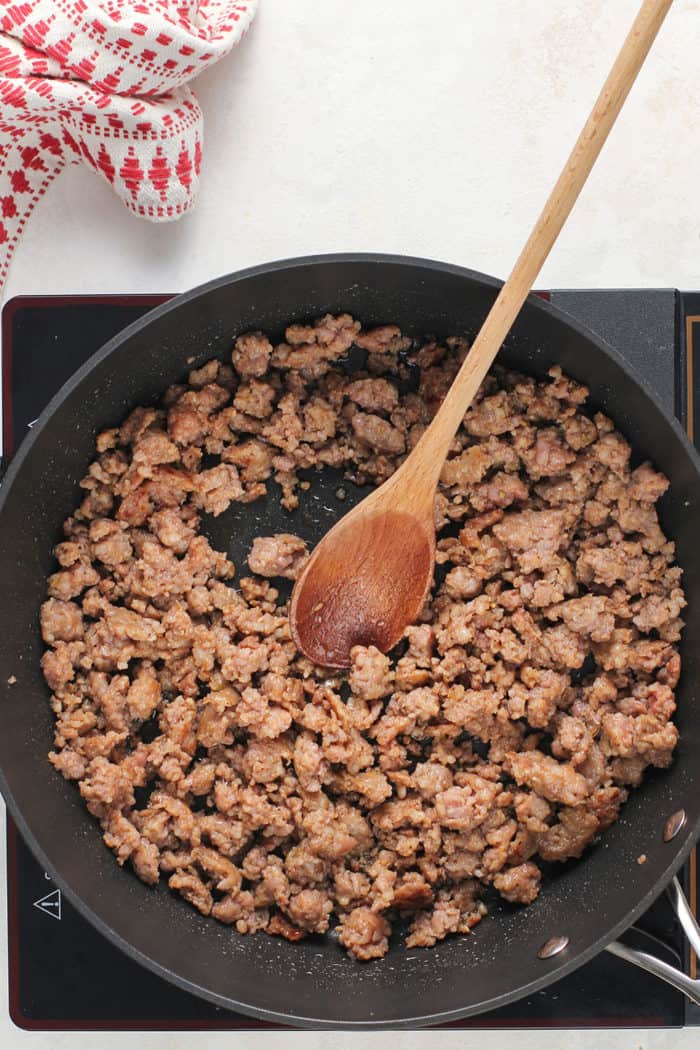 Browned, crumbled sausage in a black skillet, being stirred with a wooden spoon