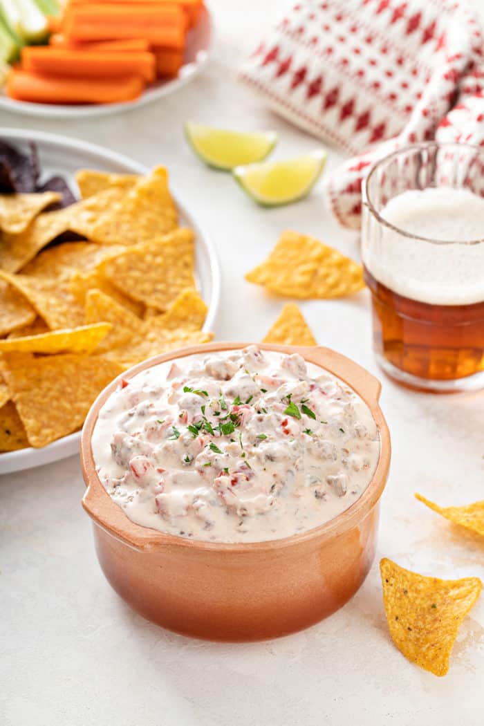 Bowl of spicy sausage dip with beer, chips, and veggies in the background