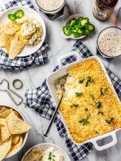 White baking dish of jalapeno popper dip with a spoon in it, next to chips and a plate of dip and chips