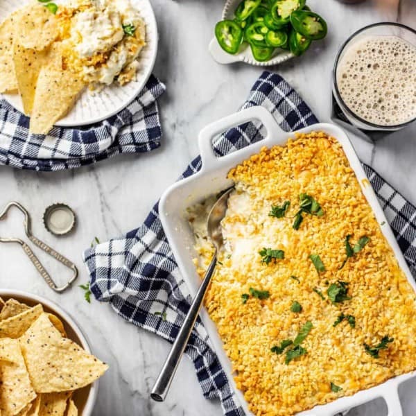 White baking dish of jalapeno popper dip with a spoon in it, next to chips and a plate of dip and chips