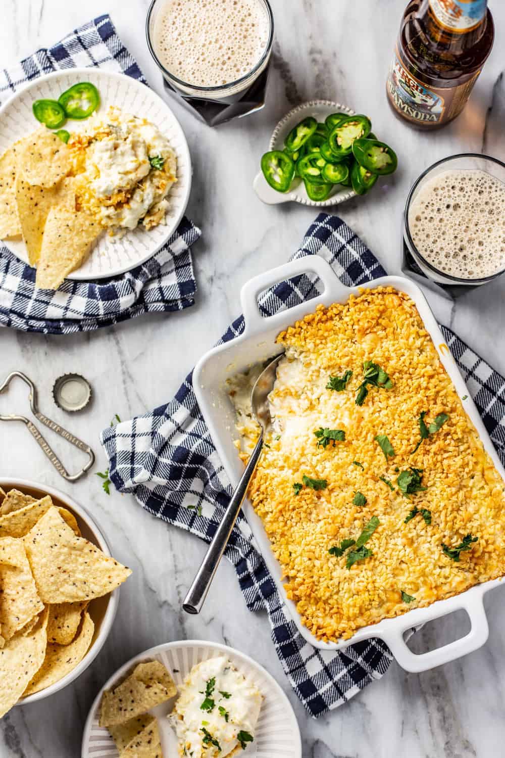 White baking dish of jalapeno popper dip with a spoon in it, next to chips and a plate of dip and chips