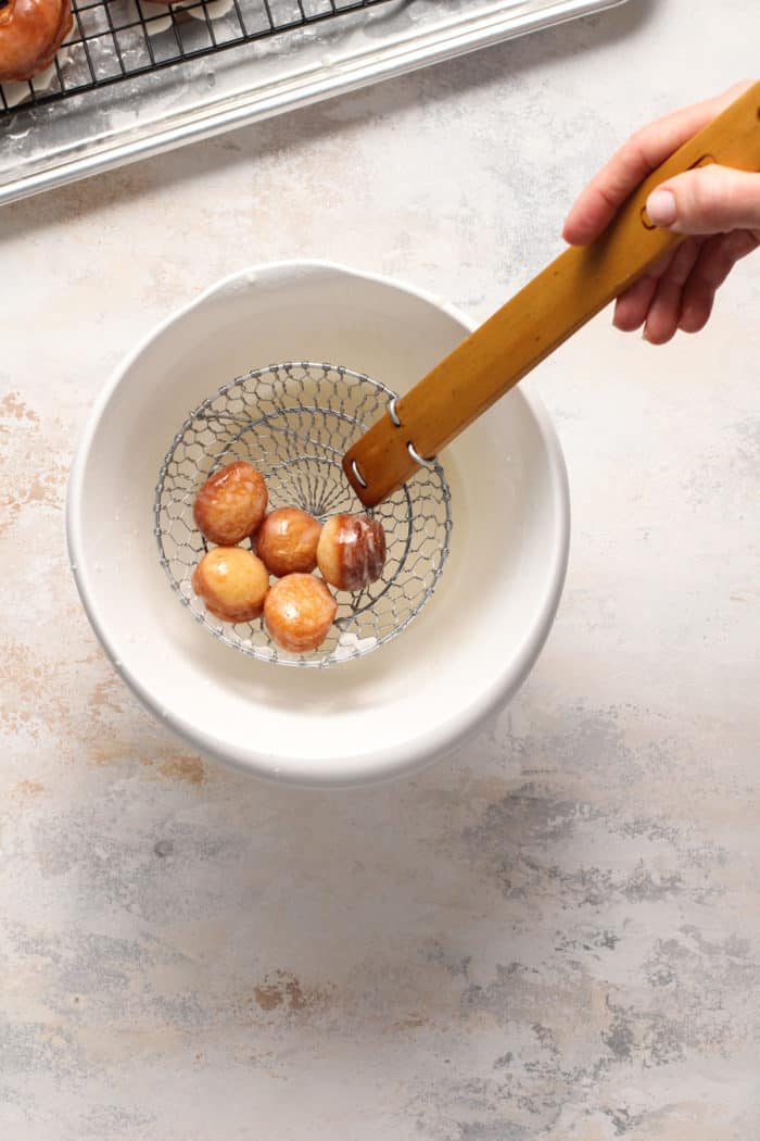 Doughnut holes being removed from a bowl of glaze with a metal spider.