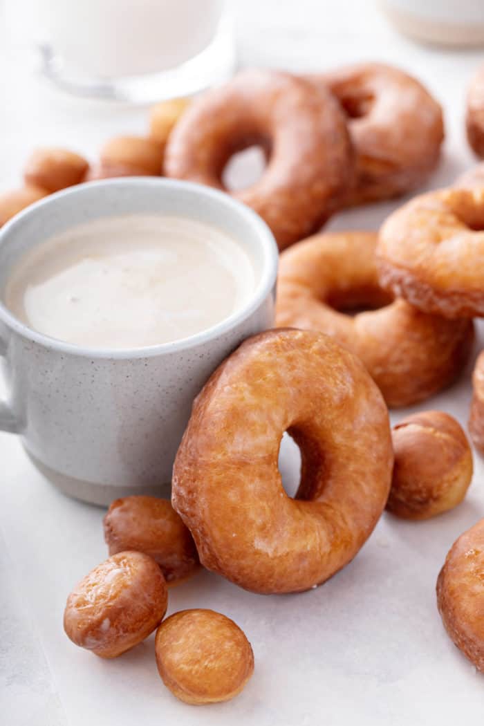 Homemade doughnuts surrounding a cup of coffee, with one of the doughnuts resting against the side of the coffee cup.