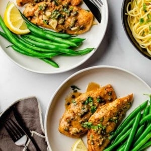 Overhead photo of two plates of Chicken Piccata served with green beans and a lemon wedge