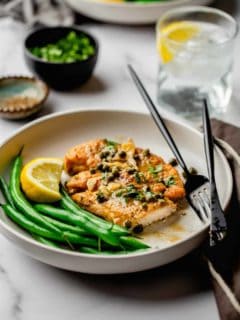 Chicken Piccata on a plate served with green beans and a lemon wedge with a black fork and knife.