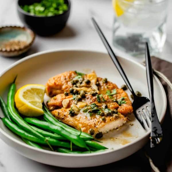 Chicken Piccata on a plate served with green beans and a lemon wedge with a black fork and knife.