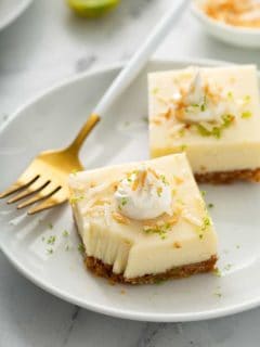 Two key lime pie bars on a white plate next to a gold fork with a white handle. The corner of one bar has a bite taken out of it