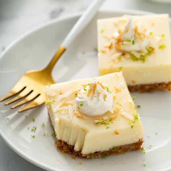 Two key lime pie bars on a white plate next to a gold fork with a white handle. The corner of one bar has a bite taken out of it