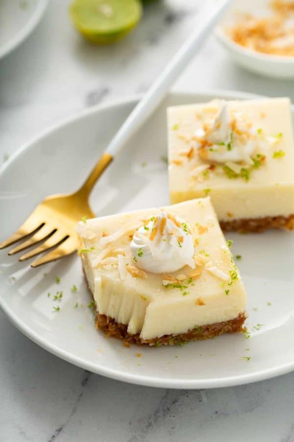 Two key lime pie bars on a white plate next to a gold fork with a white handle. The corner of one bar has a bite taken out of it