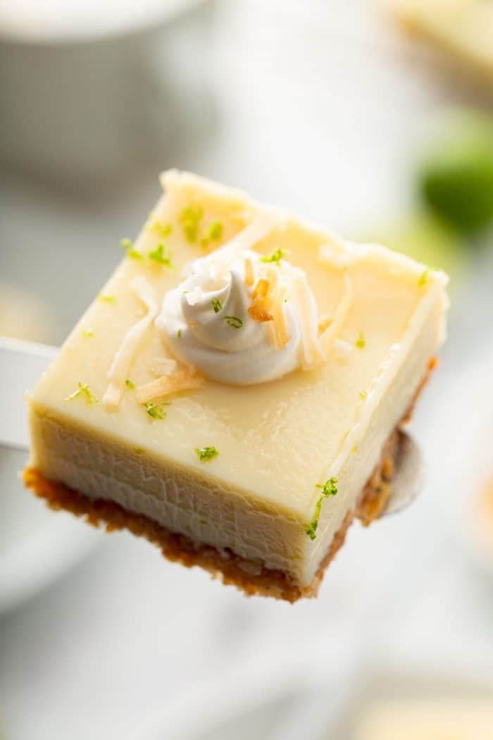 Key lime pie bar topped with whipped cream and toasted coconut being held up to the camera by an offset spatula