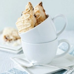 Two cherry white chocolate scones sitting in a white mug stacked on another on a plate with a spoon