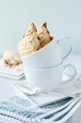 Two cherry white chocolate scones sitting in a white mug stacked on another on a plate with a spoon