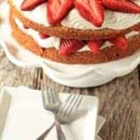 Strawberry cream cake on a cake stand behind a plate with two forks