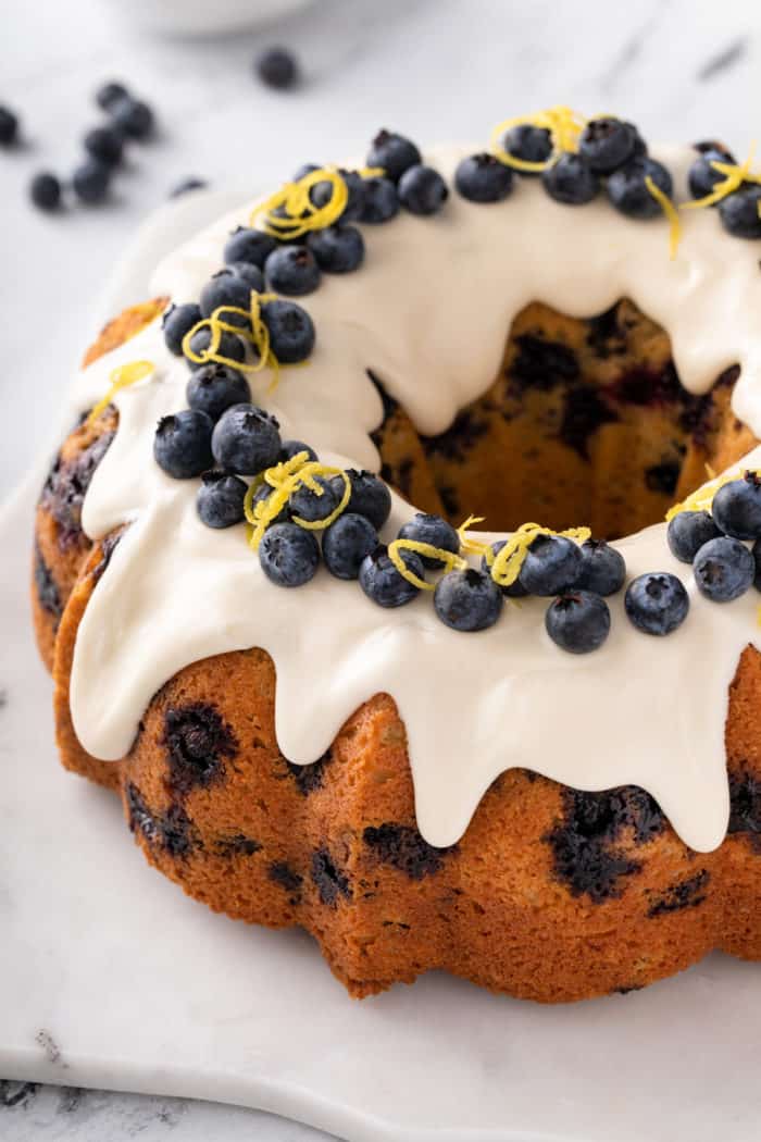 Baked lemon-blueberry bundt cake topped with cream cheese glaze and garnished with lemon zest and whole blueberries.