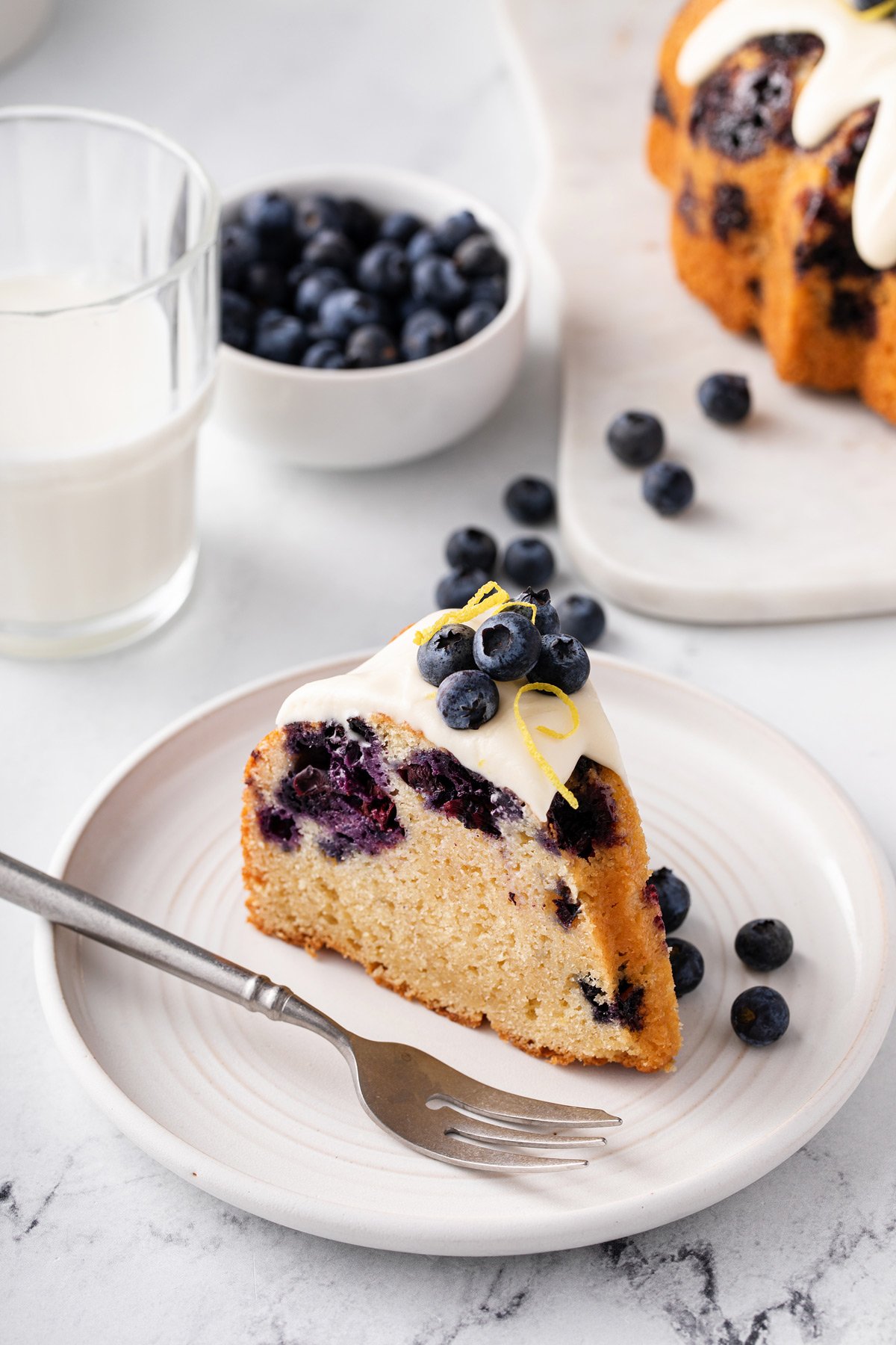 Slice of lemon-blueberry bundt cake topped with cream cheese glaze and whole blueberries set next to a fork on a white plate.