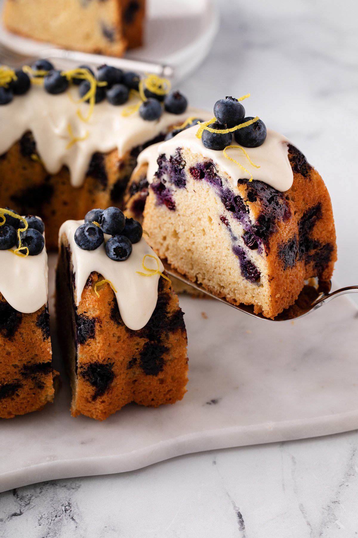 Cake server lifting a slice of cake out of a lemon-blueberry bundt cake that is topped with cream cheese glaze.
