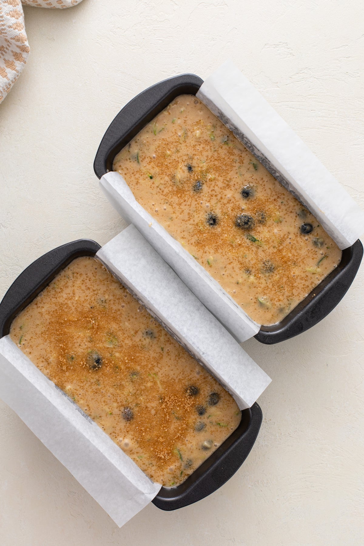 Batter for blueberry zucchini bread divided between two parchment-lined loaf pans and topped with coarse sugar.