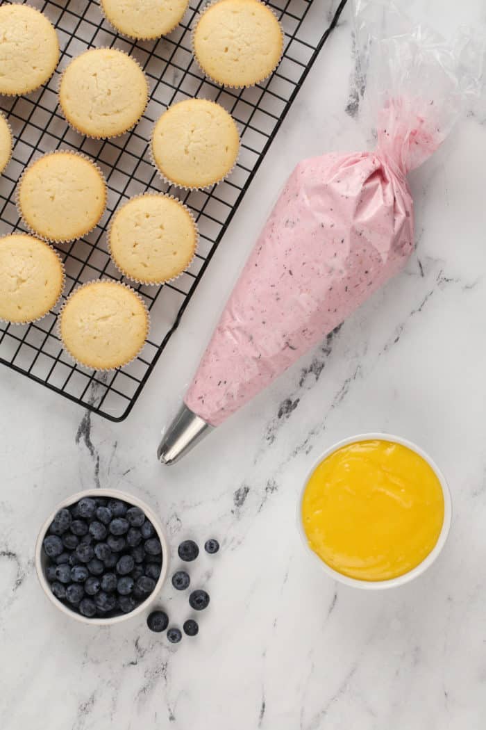 Baked lemon cupcakes on a wire rack next to a bowl of lemon curd and a piping bag filled with lemon-blueberry frosting.