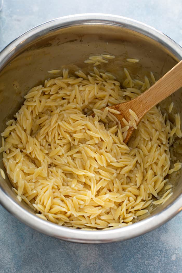 Cooked orzo pasta being stirred with a wooden spoon in a metal mixing bowl