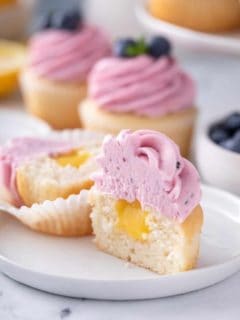 Lemon cupcake cut in half to show the lemon curd filling. The cupcake is topped with lemon-blueberry frosting.
