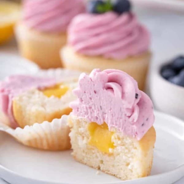 Lemon cupcake cut in half to show the lemon curd filling. The cupcake is topped with lemon-blueberry frosting.