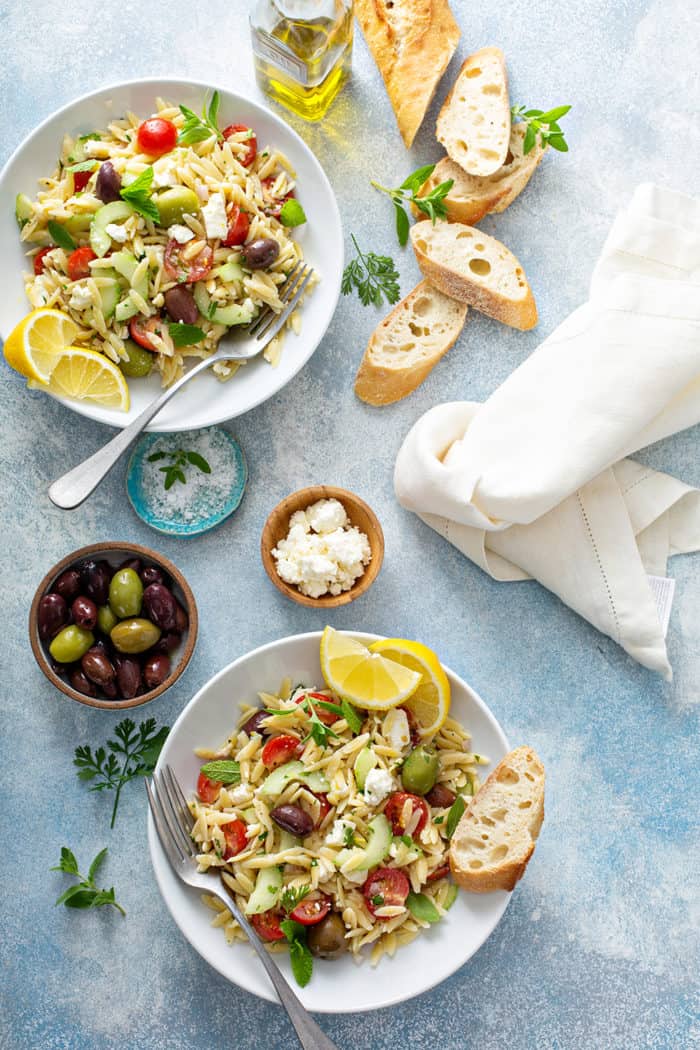 Greek orzo salad in two small white bowls surrounded by olives, feta, and slices of baguette