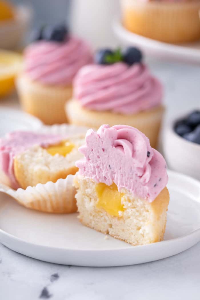 Lemon cupcake cut in half to show the lemon curd filling. The cupcake is topped with lemon-blueberry frosting.