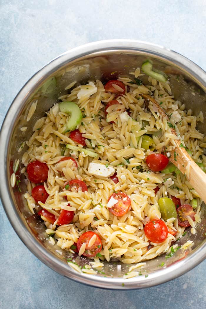 Wooden spoon stirring together greek orzo salad in a metal bowl