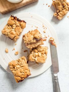 Four peanut butter and jelly bars scattered on a white plate next to a knife with peanut butter on the tip