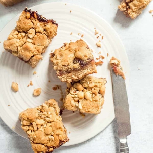 Four peanut butter and jelly bars scattered on a white plate next to a knife with peanut butter on the tip