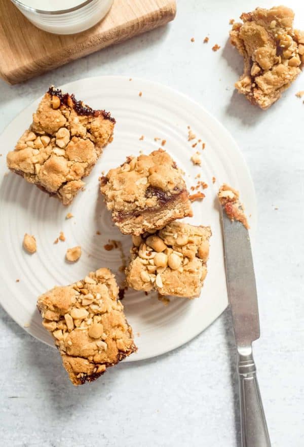 Four peanut butter and jelly bars scattered on a white plate next to a knife with peanut butter on the tip