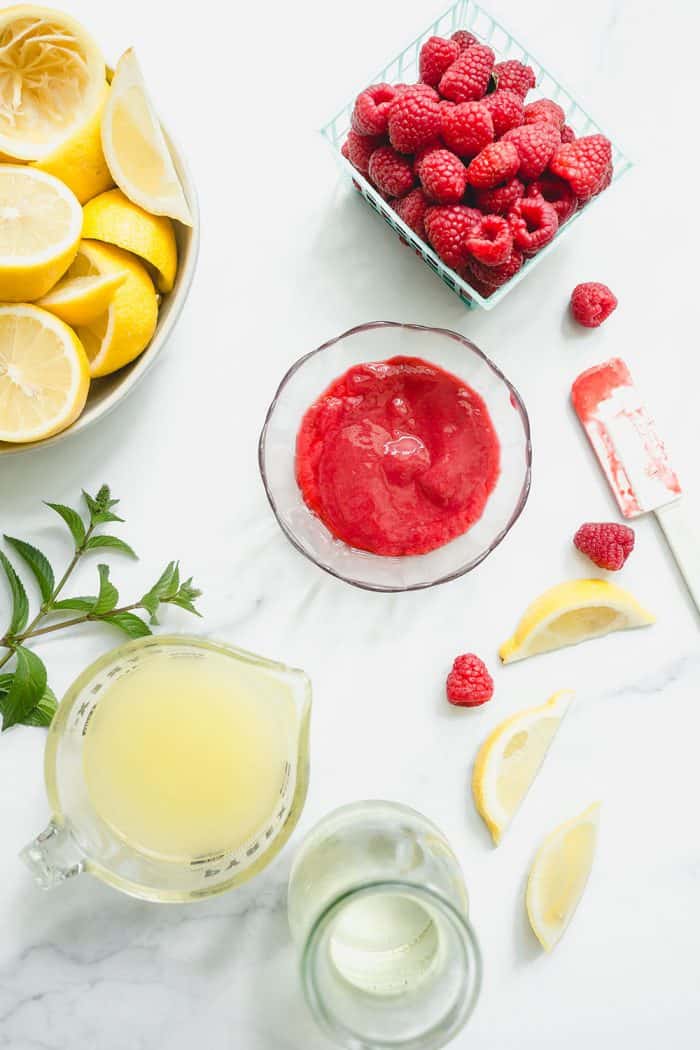 Overhead view of a bowl of raspberry puree and a measuring cup of lemon juice on a white counter, next to containers of fresh raspberries and halved lemons