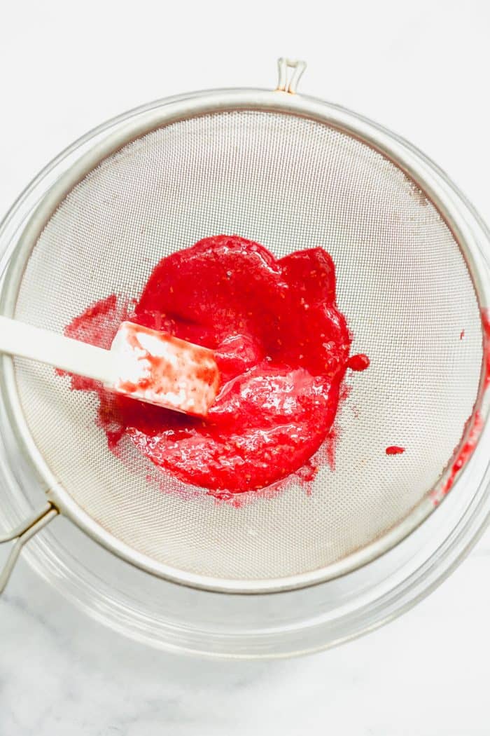 Spatula pushing raspberry puree through a fine mesh sieve set over a glass bowl on a white surface