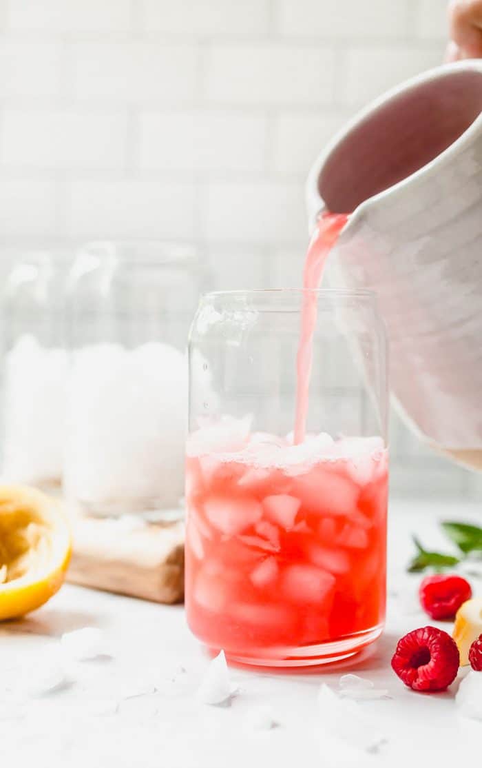 White pitcher pouring raspberry lemonade into a glass. Additional glasses with ice are in the background