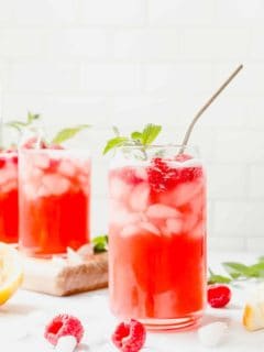 Glass of raspberry lemonade with a sprig of mint and a metal straw in the foreground, with 2 more glasses of lemonade in the background