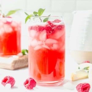 Glass of raspberry lemonade garnished with a sprig of mint on a white countertop, surrounded by fresh raspberries and another glass and pitcher of lemonade in the background