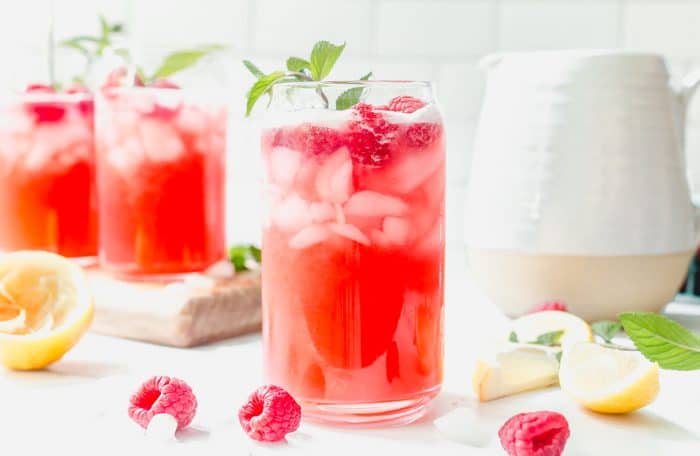 Glass of raspberry lemonade garnished with a sprig of mint on a white countertop, surrounded by fresh raspberries and another glass and pitcher of lemonade in the background