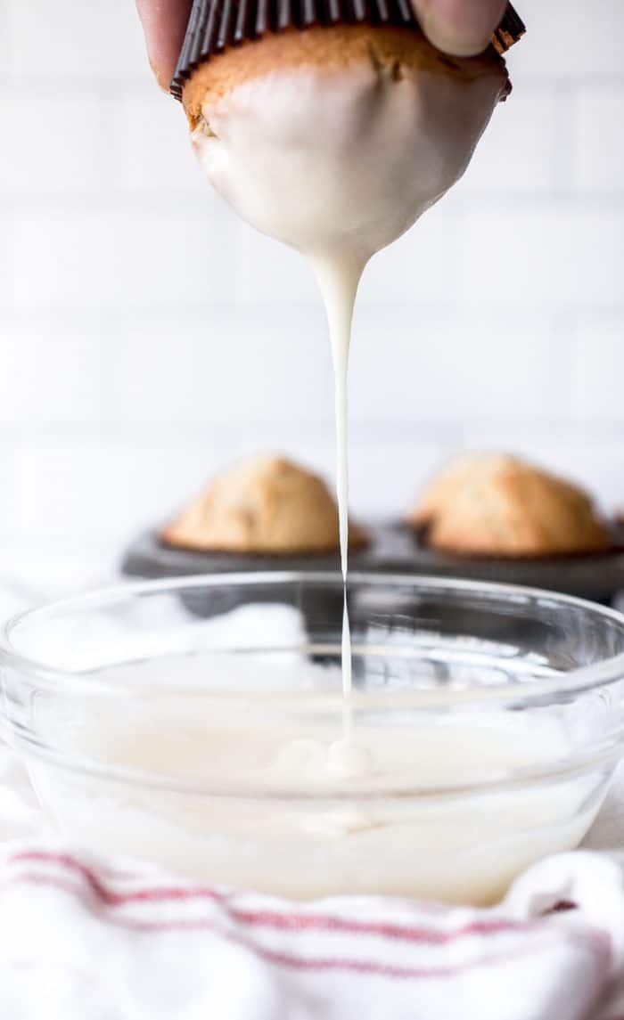 Donut muffin being lifted out of a bowl of donut glaze, with the white glaze dripping off the top of the muffin and back into the bowl