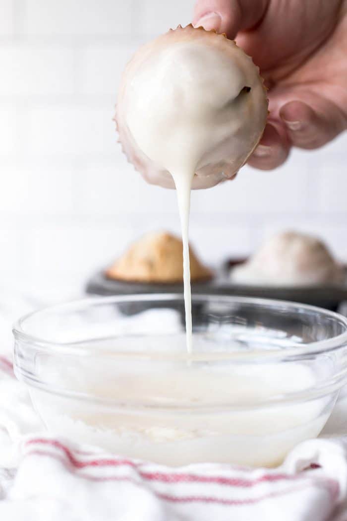 Donut muffin being lifted out of a glass bowl of donut glaze, with the excess glaze dripping back into the bowl