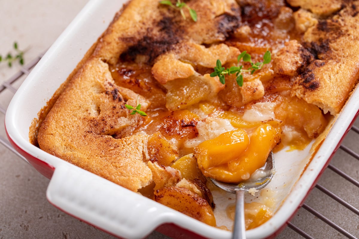 White dish filled with homemade peach cobbler. a serving spoon is dishing out a serving of the cobbler from the pan.