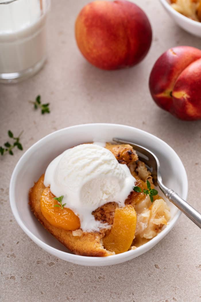 White bowl filled with a serving of homemade peach cobbler, topped with ice cream, with a spoon taking a bite of the cobbler.
