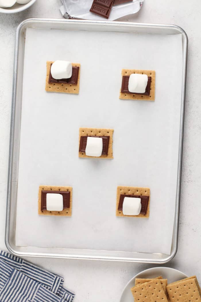 Five graham cracker squares on a parchment-lined baking sheet, each topped with a square of chocolate and a marshmallow.