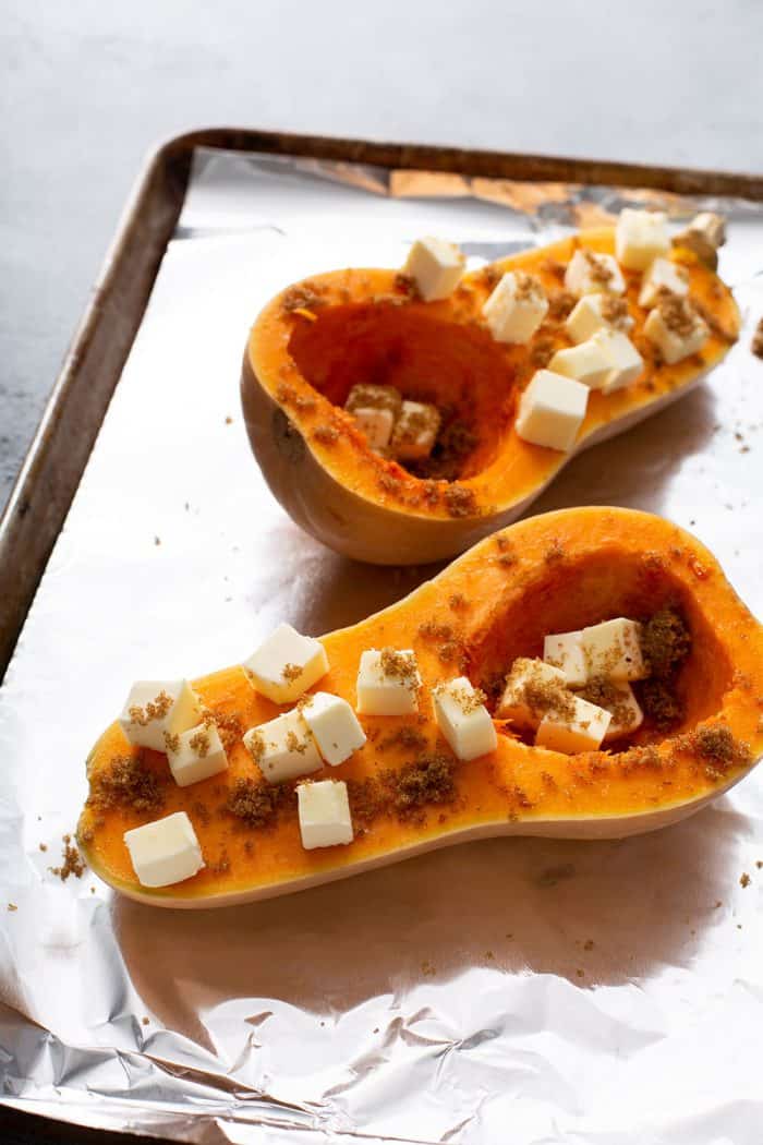 Halved butternut squash topped with butter cubes and brown sugar on a foil-lined baking sheet