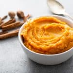 Butternut squash puree in a white bowl next to a spoon and whole cinnamon and nutmeg