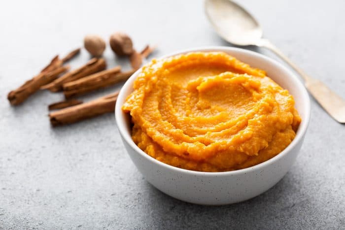 Butternut squash puree in a white bowl next to a spoon and whole cinnamon and nutmeg