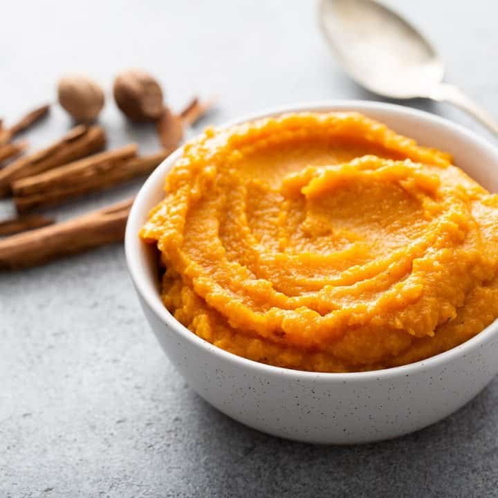 Butternut squash puree in a white bowl next to a spoon and whole cinnamon and nutmeg
