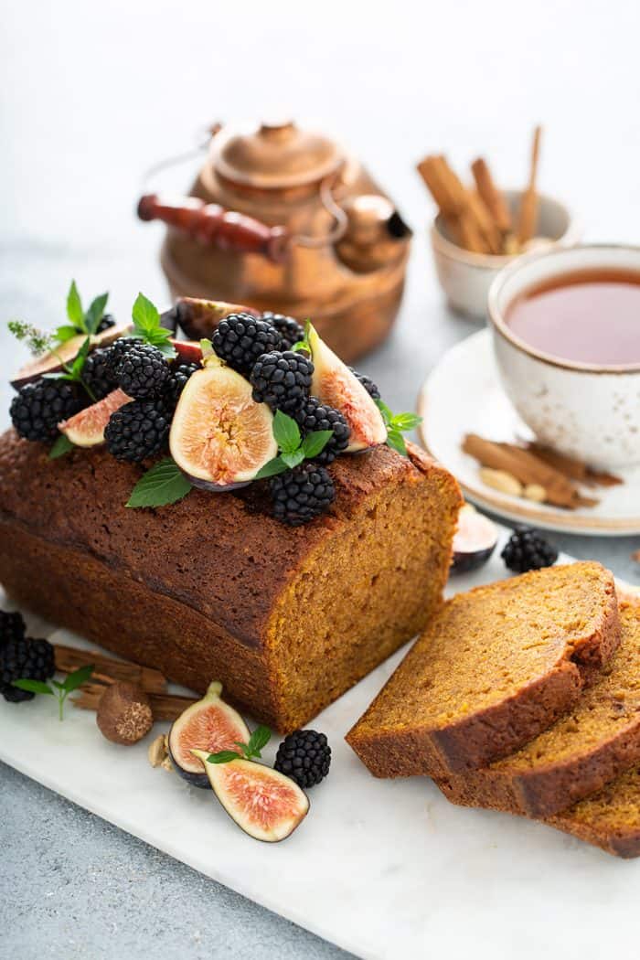 Sliced loaf of butternut squash bread topped with figs and berries on a cutting board with a teapot and cup of chai in the background