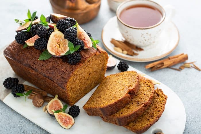 Loaf of butternut squash bread topped with figs and blackberries on a marble board with a cup of chai in the background